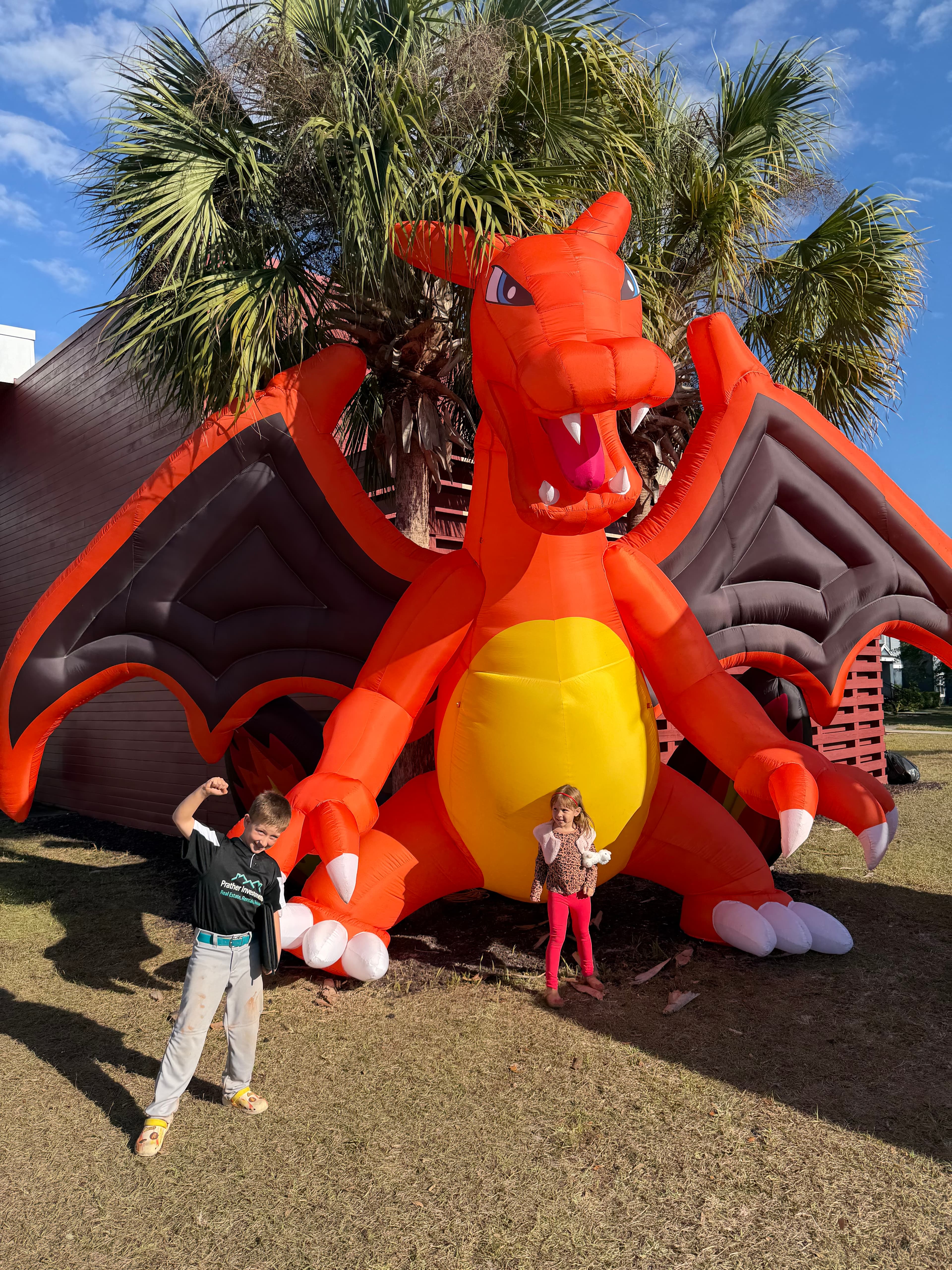 A giant inflatable Charizard towers over two children posing on a sunny lawn.