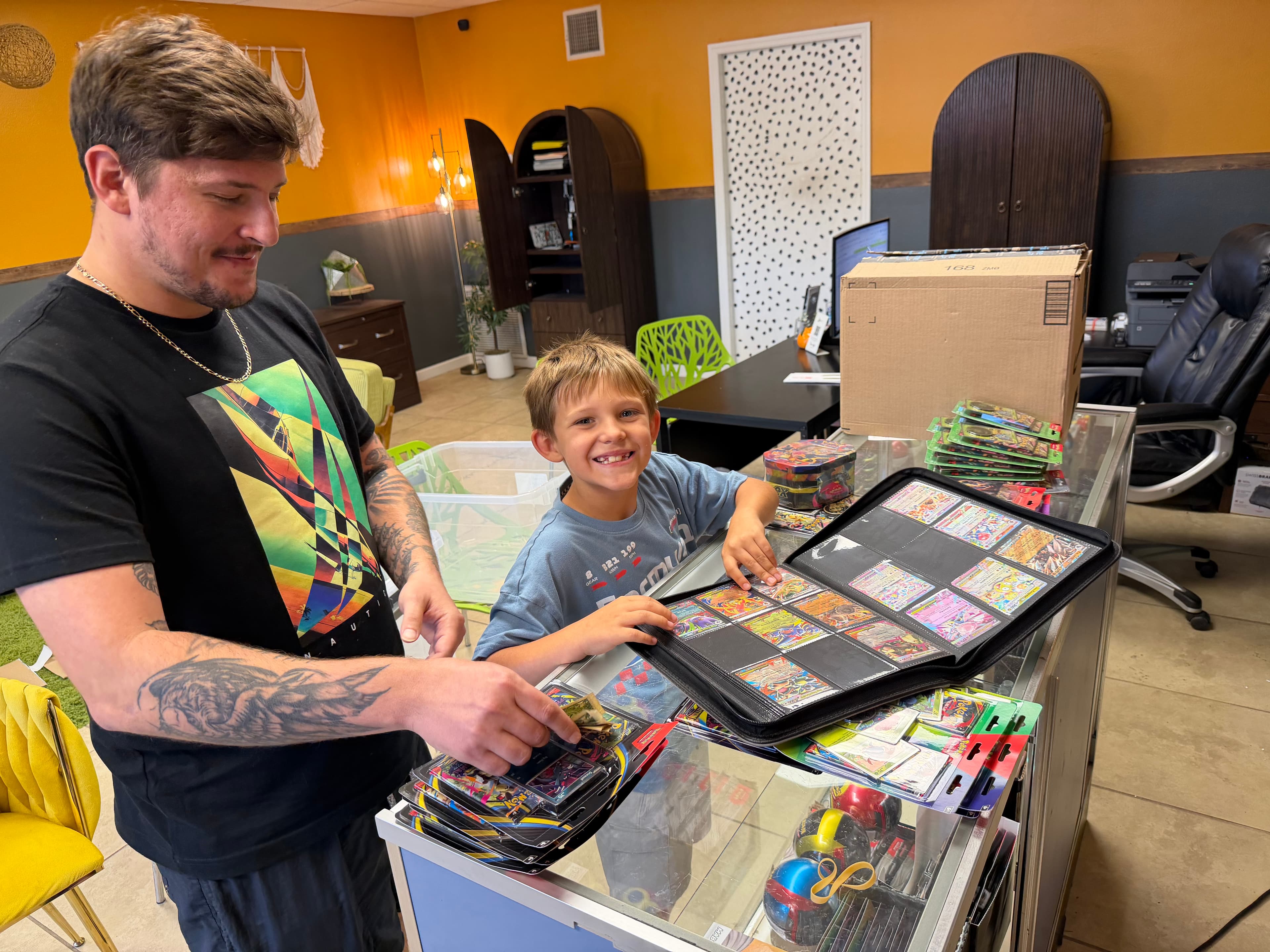 A man and a boy smile while looking at a binder of Pokémon trading cards.