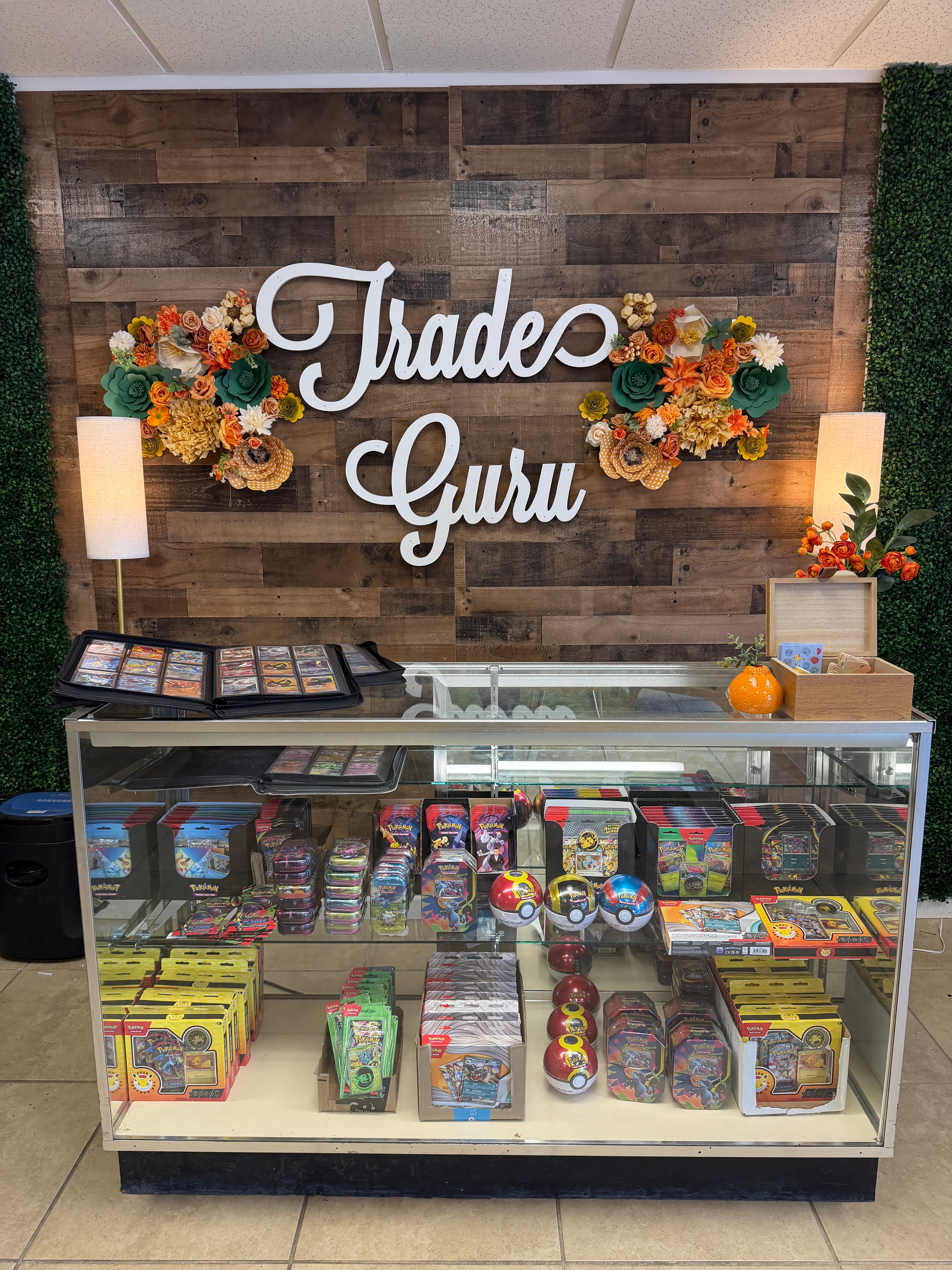 Trade Guru shop display with wooden wall, floral sign, and glass case of Pokémon products.