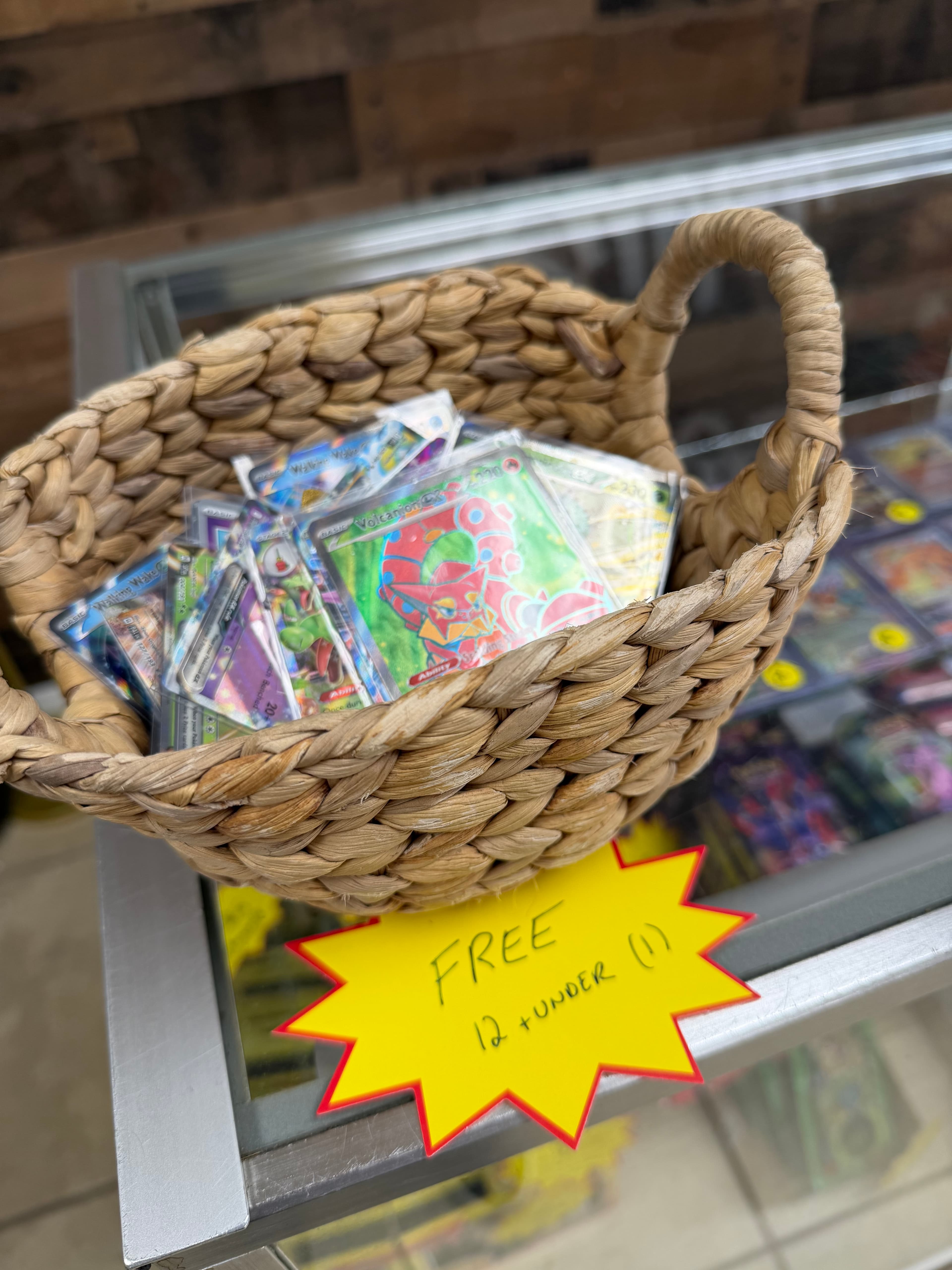 Woven basket filled with Pokémon cards on a glass counter with a yellow "Free" sign.