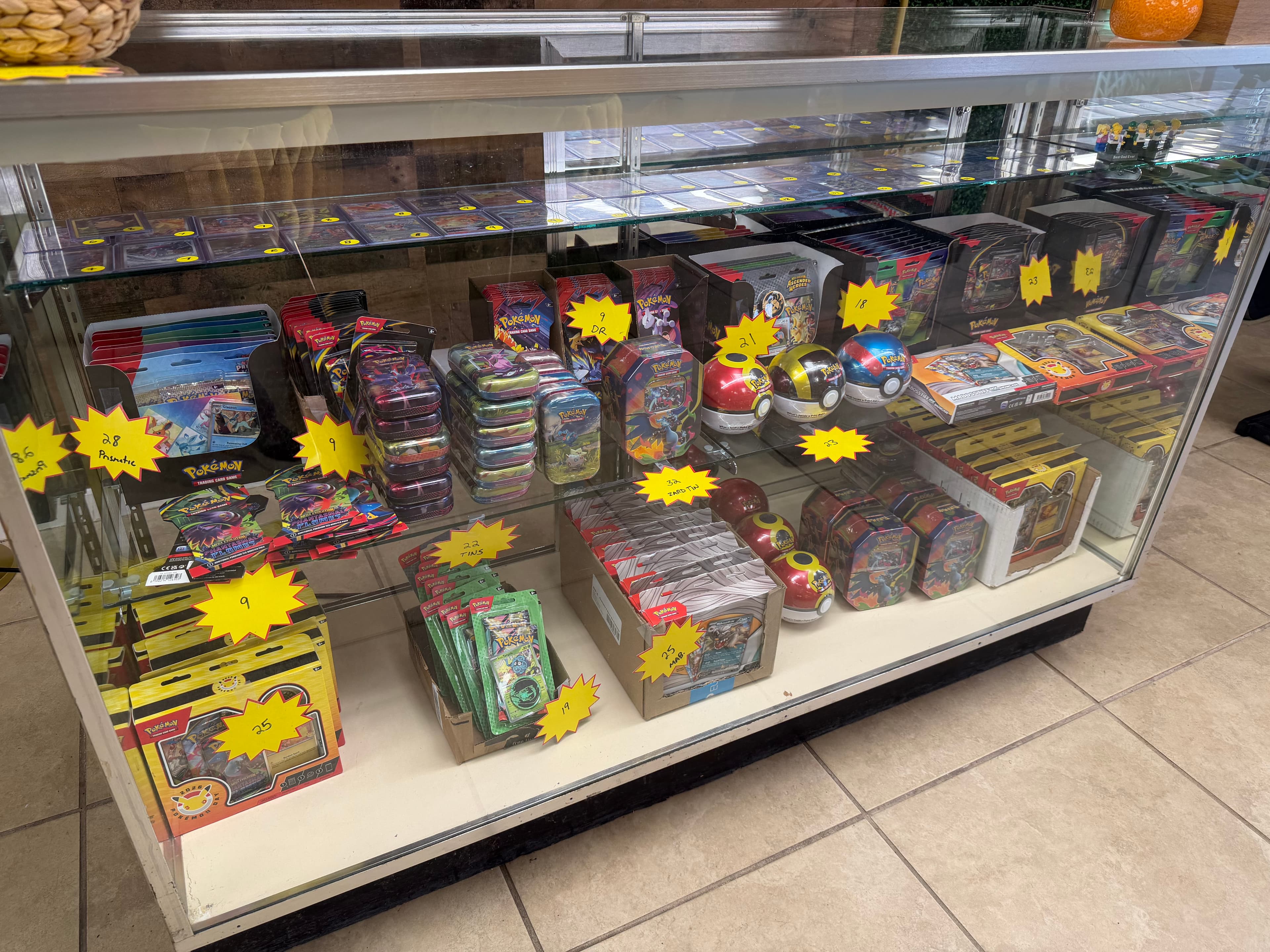 Glass display case filled with Pokémon tins, Poké Balls, card packs, and yellow price tags.