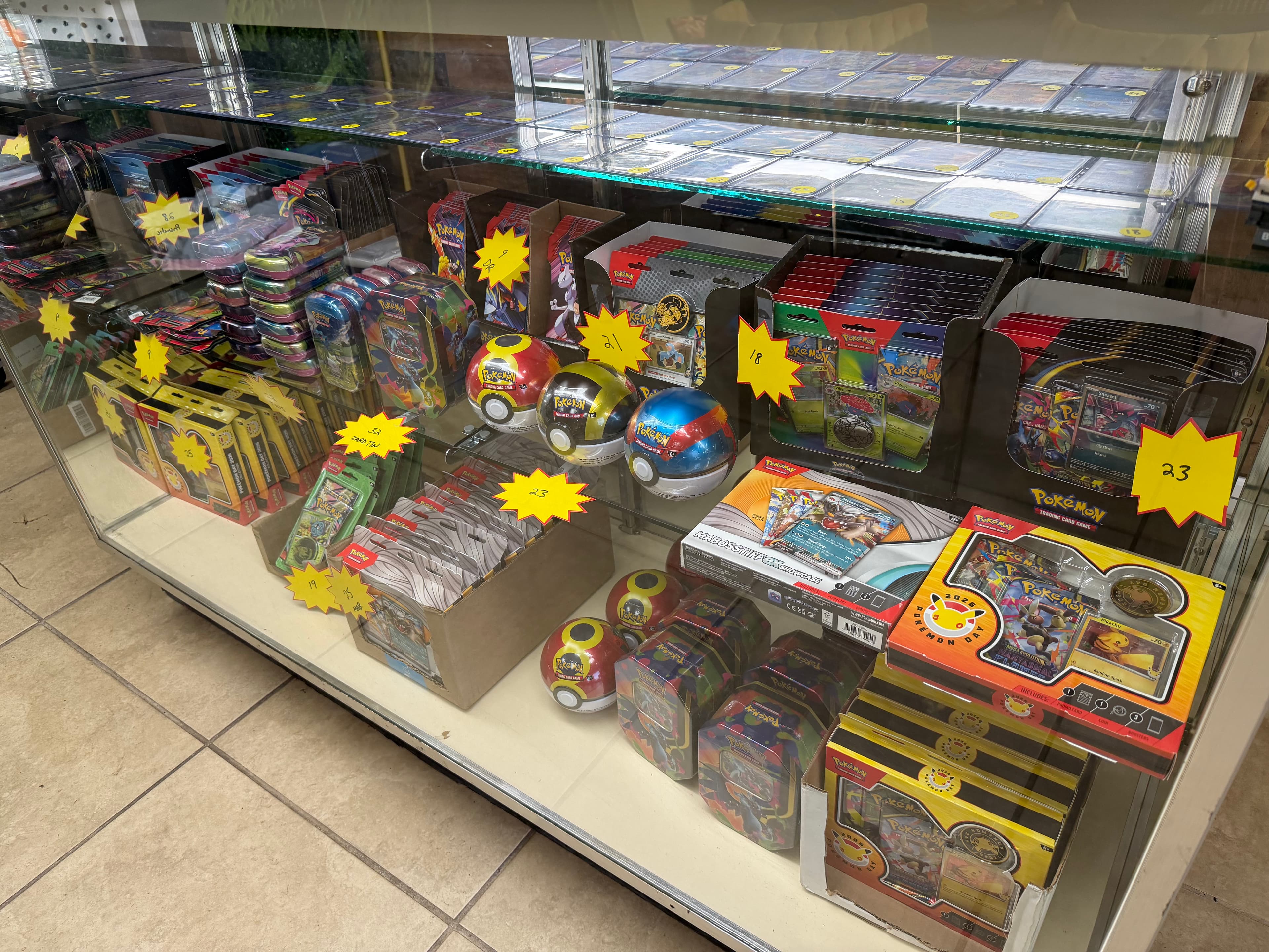 Pokémon trading card game products, including tins and Poké Balls, inside a glass display case.